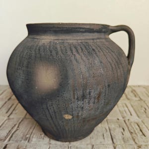 May include: A rustic, dark-colored ceramic jug with a rounded body and a single handle. The jug has a textured surface with a dark, weathered appearance, suggesting age and use. The jug is likely used for storing liquids or as a decorative piece.