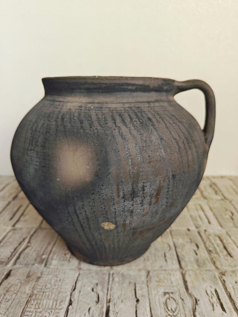 May include: A rustic, dark-colored ceramic jug with a rounded body and a single handle. The jug has a textured surface with a dark, weathered appearance, suggesting age and use. The jug is likely used for storing liquids or as a decorative piece.