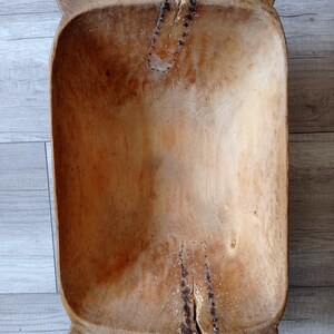 May include: A large, wooden dough bowl with a natural finish. The bowl has a rectangular shape with rounded edges and a deep well. The wood is slightly weathered and shows signs of use.