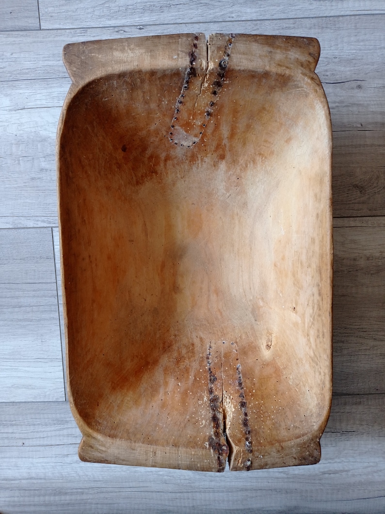 May include: A large, wooden dough bowl with a natural finish. The bowl has a rectangular shape with rounded edges and a deep well. The wood is slightly weathered and shows signs of use.