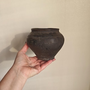 May include: A dark brown, antique-style ceramic pot with a wide, rounded body and a slightly flared rim. The pot has a rough, textured surface and appears to be handmade. It is held in a person's hand against a neutral background.