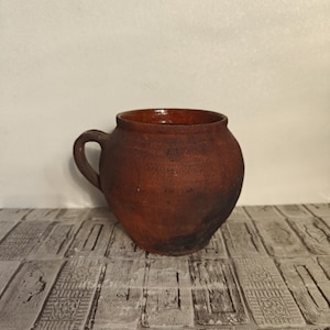 May include: A rustic, brown earthenware pot with a rounded body and a single handle. The pot has a dark, textured surface and a wide opening. The handmade pottery is likely used for cooking or storage.