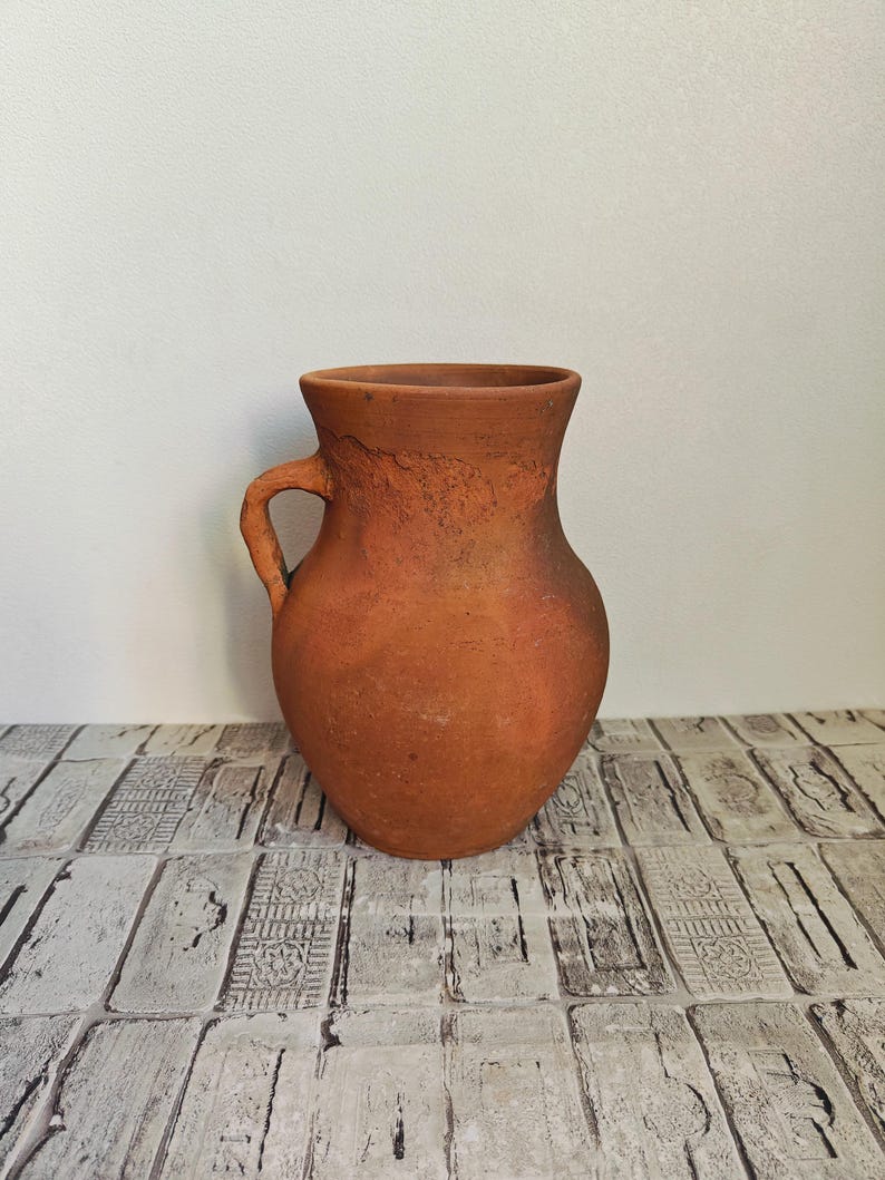 May include: A rustic, terracotta-colored pitcher with a handle. The earthenware jug has a rounded body and a slightly textured surface. The pitcher is suitable for decorative use or for serving beverages.