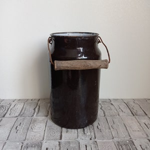 May include: A vintage brown enamel milk can with a wooden handle and rusty metal accents. The can is sitting on a tiled surface.