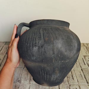 May include: A large, dark gray ceramic jug with a single handle. The jug has a rounded body and a slightly textured surface. The handle is curved and attached to the upper part of the jug. The jug is displayed on a light-colored, patterned surface.