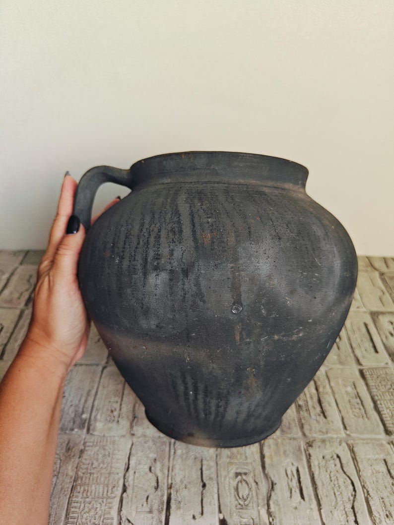 May include: A large, dark gray ceramic jug with a single handle. The jug has a rounded body and a slightly textured surface. The handle is curved and attached to the upper part of the jug. The jug is displayed on a light-colored, patterned surface.