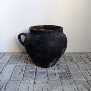 May include: A dark, rustic, handmade ceramic pot with a single handle. The pot has a rounded body and a wide opening. The exterior is a dark, matte black, with a lighter brown interior. The pot sits on a patterned surface.
