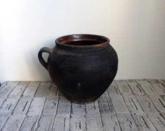 May include: A rustic, black ceramic pot with a handle. The pot has a rounded body and a dark brown interior. The exterior has a textured, matte finish. The pot is likely used for cooking or storage.