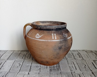 May include: A brown clay pot with a white design. The pot has a handle and is sitting on a tiled surface.