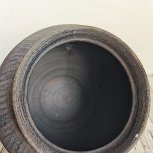 May include: A dark gray ceramic pot with a wide opening and a lid. The pot has a textured surface and a handle on the side. The interior is dark and smooth.