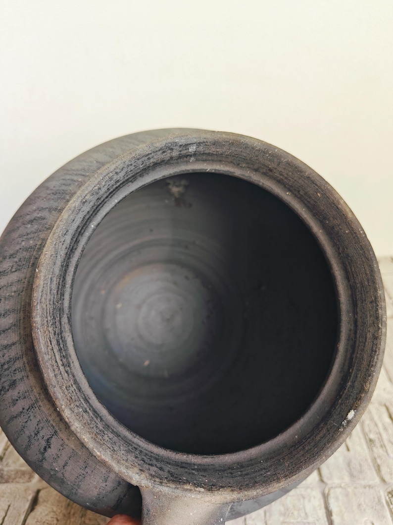 May include: A dark gray ceramic pot with a wide opening and a lid. The pot has a textured surface and a handle on the side. The interior is dark and smooth.
