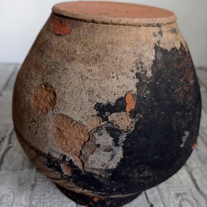 May include: A large, round, terracotta pot with a dark brown glaze. The pot is cracked and chipped, showing the terracotta underneath. The pot is sitting on a white surface.