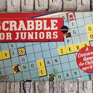 May include: A red and blue board game box with the title "SCRABBLE FOR JUNIORS" and a blue and yellow game board with letters on it. The box also includes a red circle with the text "A Crossword Game for Children ages 6 to 10".