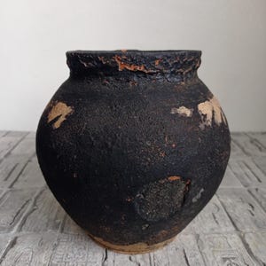 May include: A black clay pot with a rounded body and a slightly flared rim. The pot has a rustic, aged appearance with visible cracks and chips in the glaze.