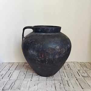 May include: A dark charcoal-colored ceramic vessel with a rounded body and a single handle on the left side. The surface features a weathered, matte texture with subtle brown and rust-colored patches visible across the exterior of the pot.