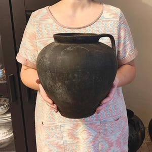 May include: A large, round, matte black ceramic vessel with a wide opening, a thick rim, and a single curved handle on the side. The surface of the pot features a weathered, textured finish with subtle gray variations and a faint, thin white line.