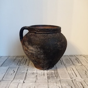 Puede incluir: Una olla de cerámica marrón oscuro de estilo antiguo con un solo asa. La olla tiene un cuerpo redondeado y un borde ligeramente ensanchado. El exterior tiene un aspecto texturizado y desgastado, lo que sugiere edad y uso. La olla probablemente se utiliza para almacenamiento o exhibición.