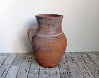 May include: A terracotta pitcher with a handle. The pitcher is brown and has a white glaze. It is sitting on a gray tiled surface.