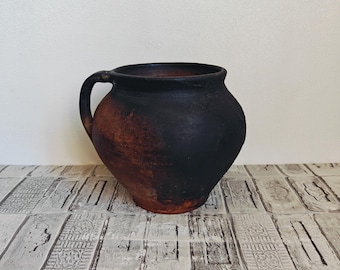 May include: A rustic, dark brown ceramic jug with a handle. The jug has a rounded body and a slightly flared rim. The exterior shows a gradient of colors, from black to reddish-brown. The jug is likely handmade and has a vintage aesthetic.