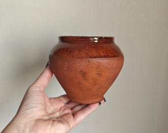 Puede incluir: Una pequeña vasija de barro color terracota con un cuerpo ancho y cónico, y un borde liso esmaltado en marrón oscuro. La parte inferior del recipiente presenta un acabado mate, texturizado y terroso con variaciones visibles en la superficie.