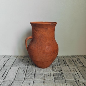Może przedstawiać: Rustykalny dzban w kolorze terakoty z jednym uchwytem. Naczynie gliniane ma pękaty korpus i lekko rozszerzoną krawędź. Powierzchnia ma teksturowany, postarzany wygląd, sugerujący, że jest ręcznie robiony. Dzban stoi na szarej i białej wzorzystej powierzchni.