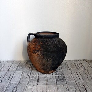 May include: A large, dark brown, earthenware pot with a single handle. The pot has a rough, textured surface and appears to be very old.