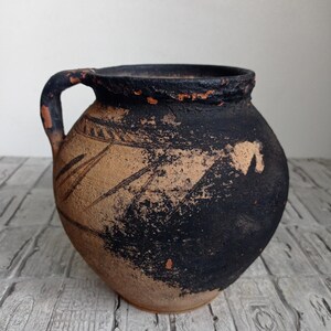 May include: A brown clay pot with a black glaze and a single handle. The pot has a rustic, aged appearance and is decorated with simple, hand-drawn lines.