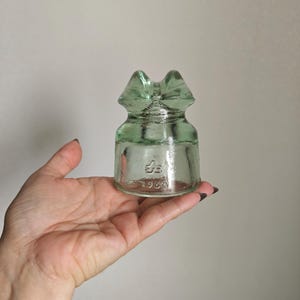 Op de afbeelding: Een lichtgroene glazen elektrische isolator met een cilindrische basis en een ingekeepte bovenkant. Het glas is voorzien van reliëfmarkeringen, waaronder een symbool en het getal 1966. Het object is doorschijnend met een glad, gegoten oppervlak.