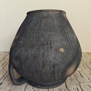 May include: A dark gray, handcrafted ceramic vase with a rounded body and a single handle. The vase has a textured surface with vertical lines and a slightly flared rim. The vase is sitting on a wooden surface.