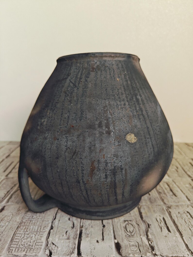 May include: A dark gray, handcrafted ceramic vase with a rounded body and a single handle. The vase has a textured surface with vertical lines and a slightly flared rim. The vase is sitting on a wooden surface.