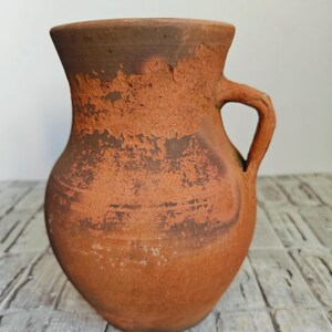 May include: A rustic, terracotta-colored pitcher with a handle. The jug has a wide base that tapers to a narrower neck and a flared rim. The surface has a textured, aged appearance with darker brown patches.