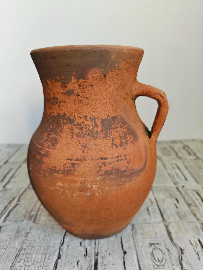 May include: A rustic, terracotta-colored pitcher with a handle. The jug has a wide base that tapers to a narrower neck and a flared rim. The surface has a textured, aged appearance with darker brown patches.