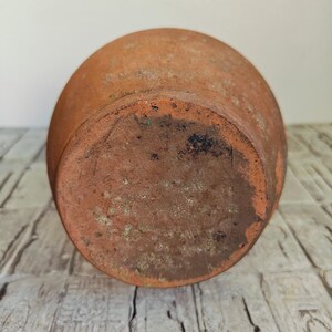 May include: A weathered, terracotta-colored ceramic pot, viewed from the bottom. The pot has a rounded shape with a slightly textured surface. The base shows signs of age and use, with darker areas and some discoloration.