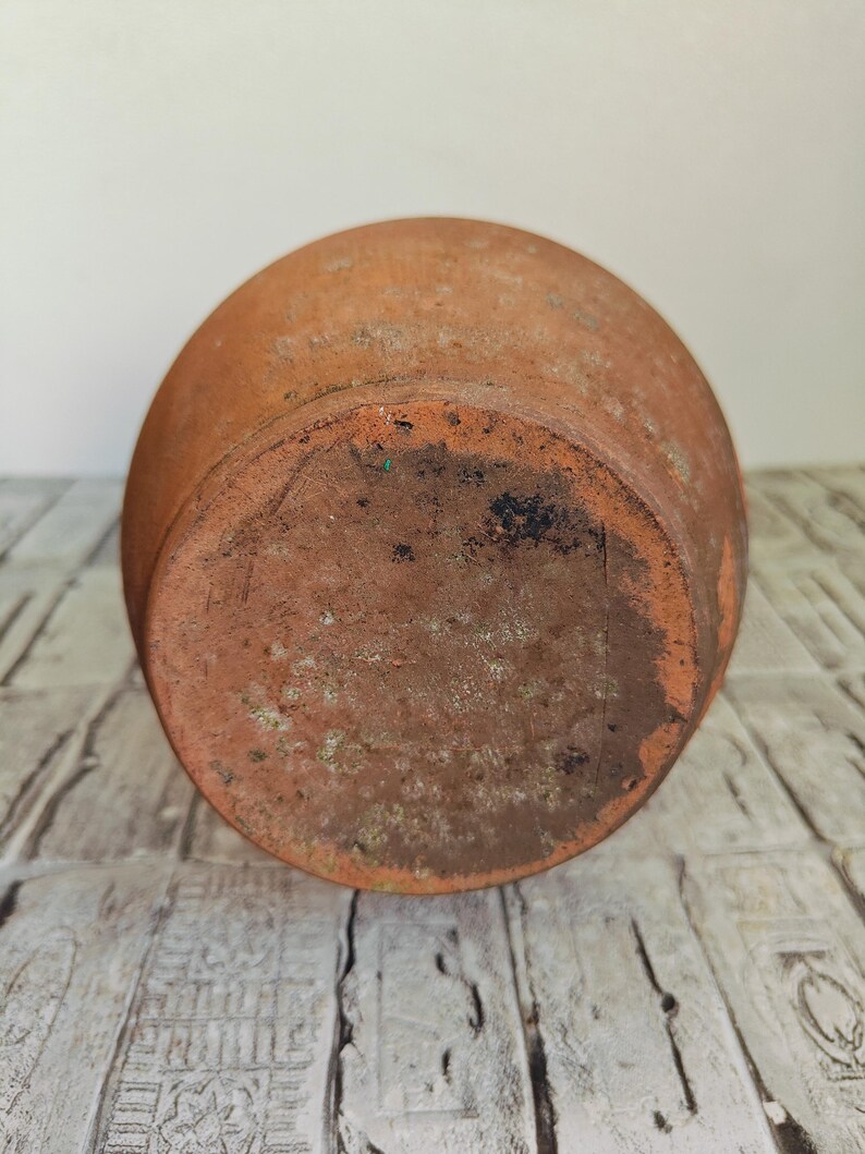 May include: A weathered, terracotta-colored ceramic pot, viewed from the bottom. The pot has a rounded shape with a slightly textured surface. The base shows signs of age and use, with darker areas and some discoloration.