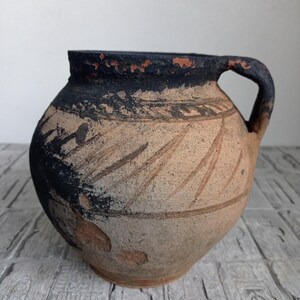 May include: A brown clay pot with a black rim and a single handle. The pot has a decorative design of lines and swirls.