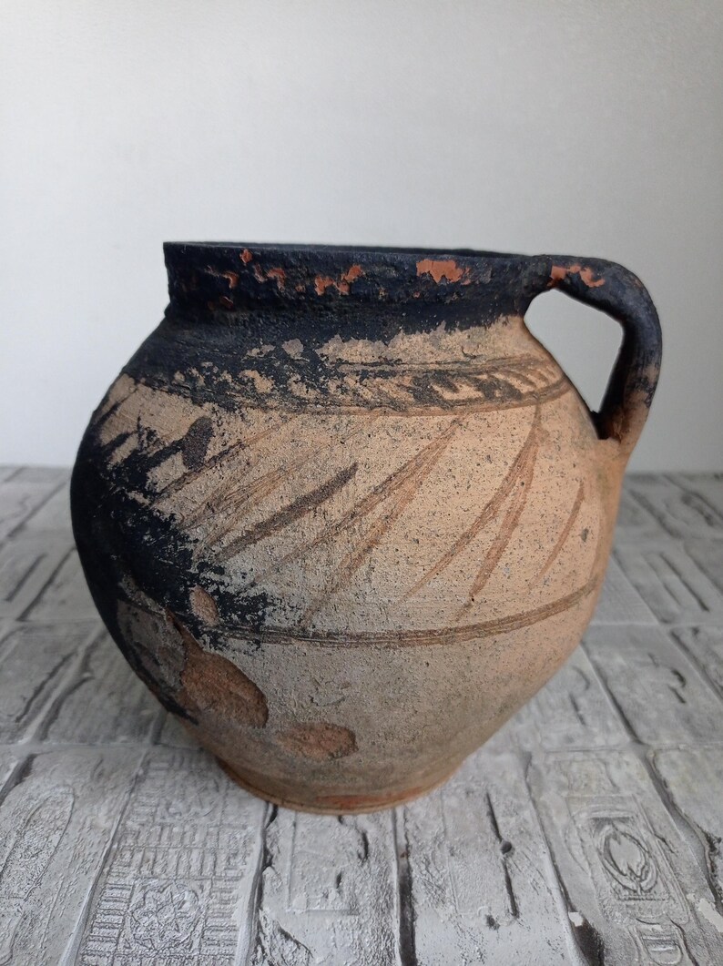 May include: A brown clay pot with a black rim and a single handle. The pot has a decorative design of lines and swirls.