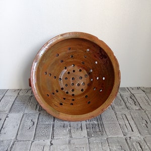 May include: A terracotta colander with a brown glaze and a circular shape. The colander has many small holes throughout the bottom.