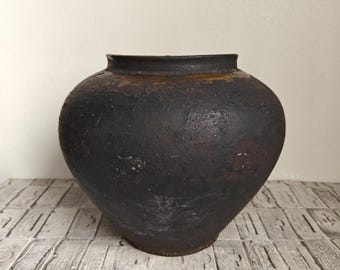 Puede incluir: Un recipiente de cerámica oscuro, redondo y bulboso con un acabado mate negro y carbón fuertemente texturizado. La vasija presenta una boca ancha con un borde ligeramente acampanado y una base cónica, mostrando un aspecto rústico y desgastado.