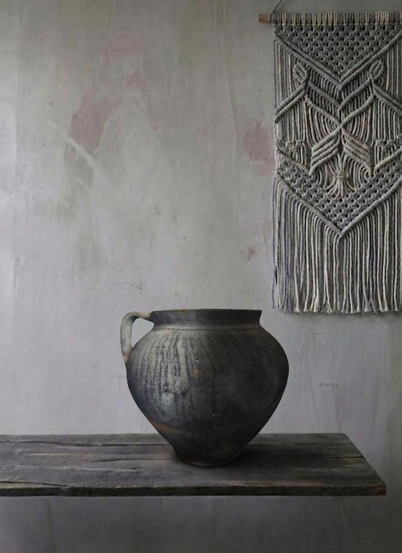 May include: A dark, aged ceramic pot with a single handle sits on a weathered wooden shelf. The pot has a rounded body and a dark, textured surface. A macrame wall hanging with a geometric design hangs in the background.