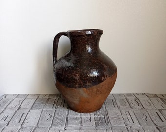 Puede incluir: Una jarra grande de estilo antiguo con asa. La parte superior es de un marrón oscuro moteado, que pasa a una base de color terracota más clara. La jarra tiene un cuerpo redondeado y un cuello estrecho, lo que sugiere un uso decorativo.