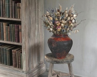 Pode incluir: Um vaso de cerâmica arredondado e escuro com uma secção superior castanho-avermelhada e uma base preta, preenchido com um arranjo de flores secas, vagens e ervas, colocado num pequeno banco de madeira desgastada com três pernas.