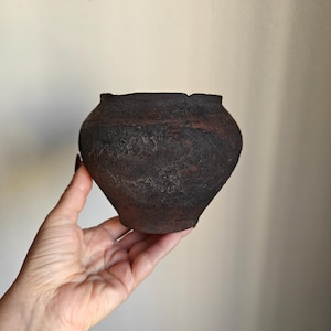 Może przedstawiać: Mały, okrągły, ciemnobrązowy ceramiczny garnek o szorstkiej, teksturowanej powierzchni i szerokim otworze. Naczynie ma pękaty korpus zwężający się ku wąskiej podstawie, wykończony matową, sprawiającą wrażenie zwietrzałej powłoką o zmiennych odcieniach.