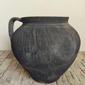 May include: A dark, rustic, handmade ceramic pot with a single handle. The pot has a rounded body and a wide opening. The exterior is a dark charcoal color with visible texture, suggesting a traditional pottery technique. The pot sits on a light-colored, textured surface.