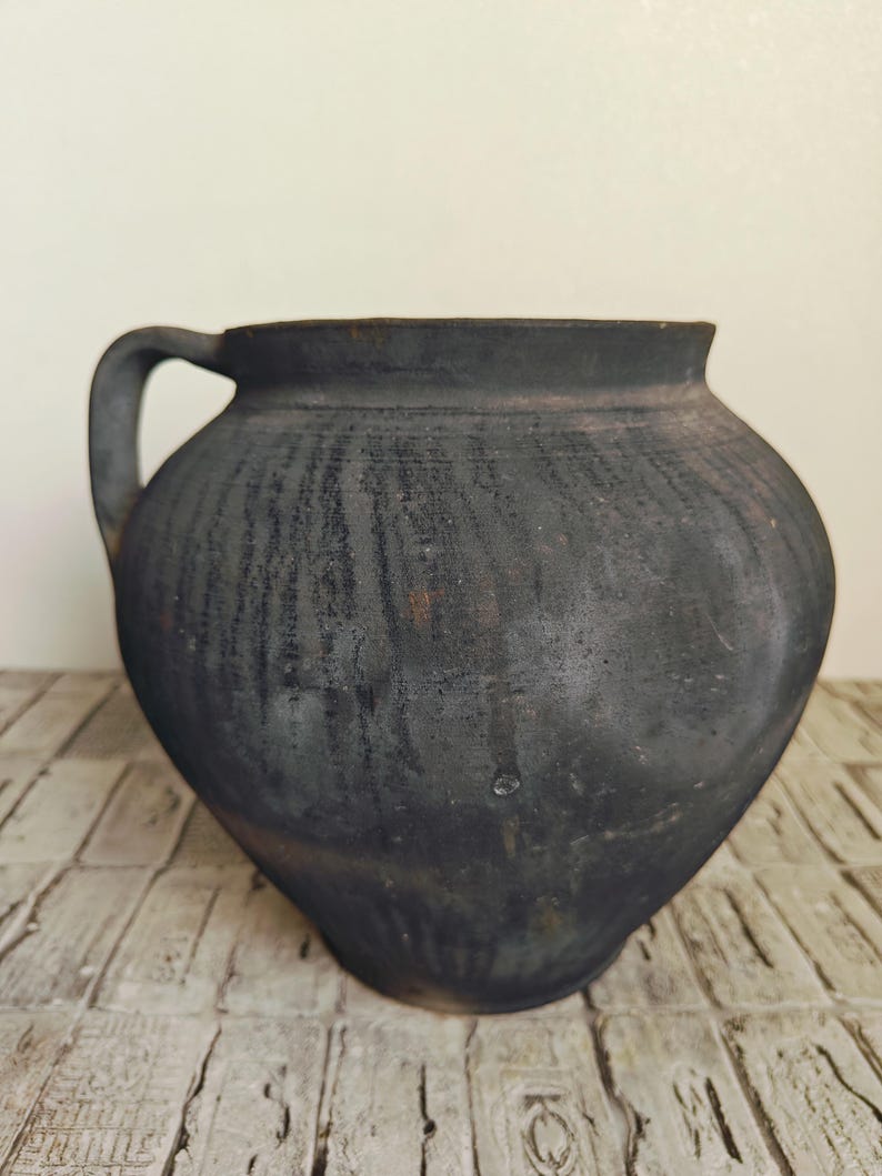 May include: A dark, rustic, handmade ceramic pot with a single handle. The pot has a rounded body and a wide opening. The exterior is a dark charcoal color with visible texture, suggesting a traditional pottery technique. The pot sits on a light-colored, textured surface.