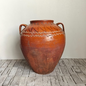 Puede incluir: Una jarra grande y redonda de barro en color terracota con dos asas laterales. La sección superior presenta un acabado brillante con patrones de líneas onduladas en blanco y marrón oscuro pintados a mano alrededor del cuello.