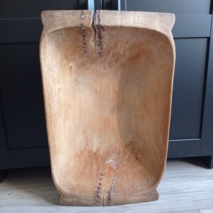 May include: A large, wooden dough bowl with a natural wood finish. The bowl is rectangular in shape and has a slight curve to the sides. The bowl is made of a single piece of wood and has a few cracks and imperfections.