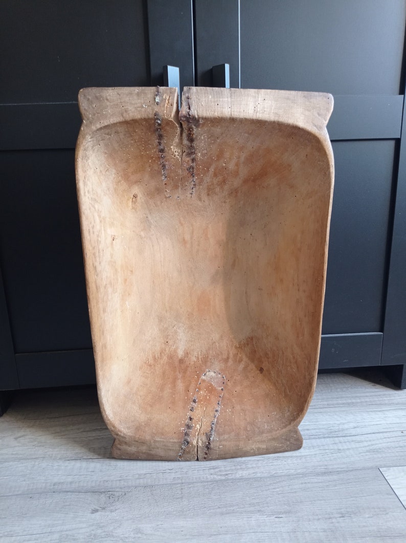 May include: A large, wooden dough bowl with a natural wood finish. The bowl is rectangular in shape and has a slight curve to the sides. The bowl is made of a single piece of wood and has a few cracks and imperfections.