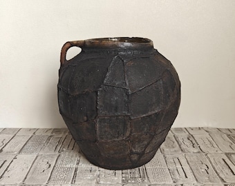 Op de afbeelding: Een donkerbruine, ronde keramische pot met een textuur die lijkt op een patchworkpatroon. De pot heeft één handvat aan de bovenzijde en een brede opening met een gladde, donkere rand. De buitenkant heeft een matte en verweerde uitstraling.