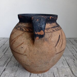 May include: A brown clay pot with a black rim and handle. The pot has a geometric design etched into the surface.