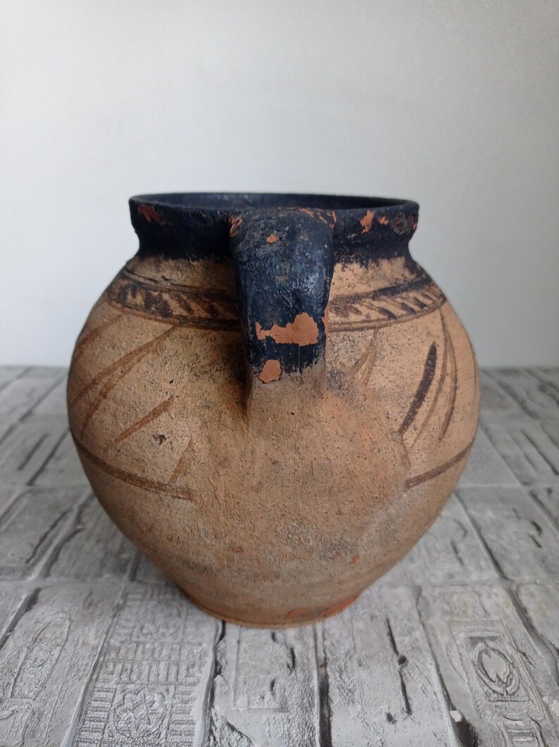 May include: A brown clay pot with a black rim and handle. The pot has a geometric design etched into the surface.